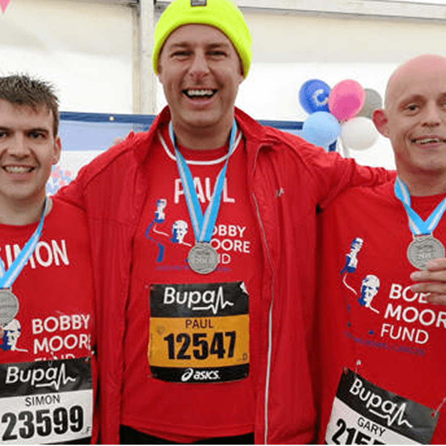 A photo of three men after a race wearing Bobby Moore Fund for Cancer Research UK shirts looking happy.