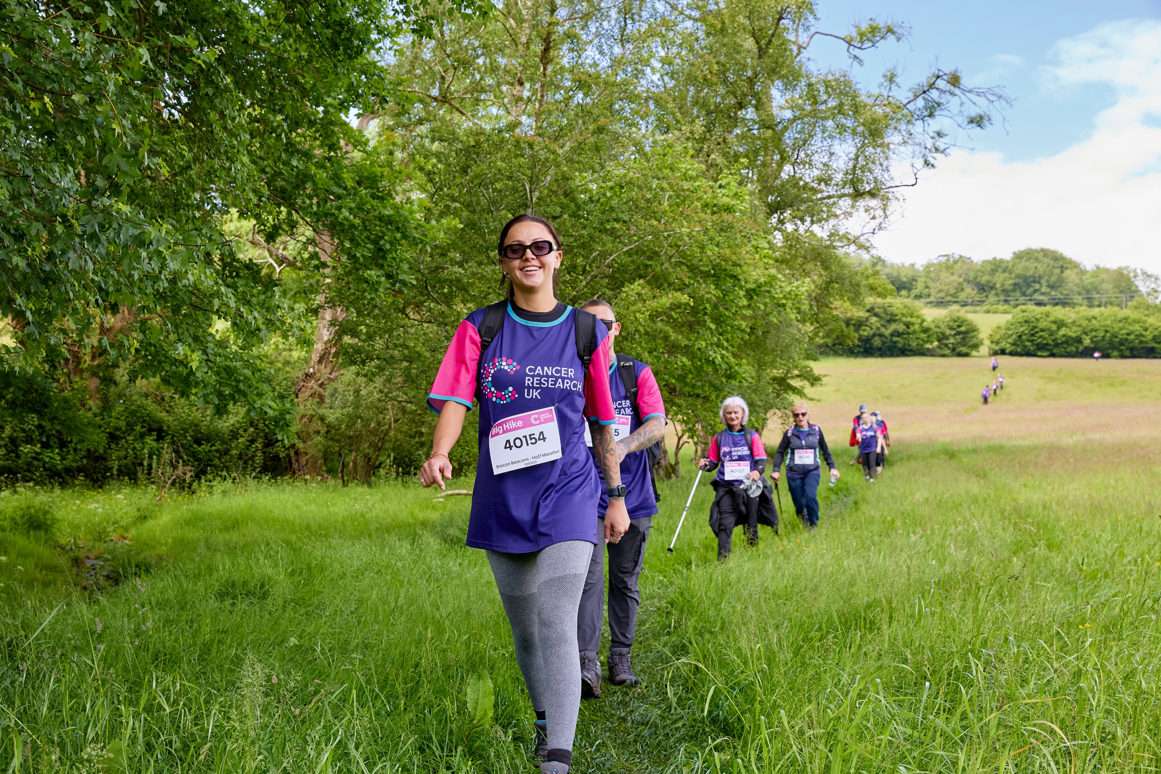 A Big Hike participant in Cancer Research UK shirts walks on a grassy path, with other hikers following behind her.