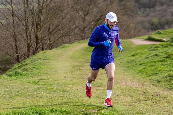 A runner in CRUK outfit and a white CRUK cap runs up a grassy path on a hill with trees in the background.