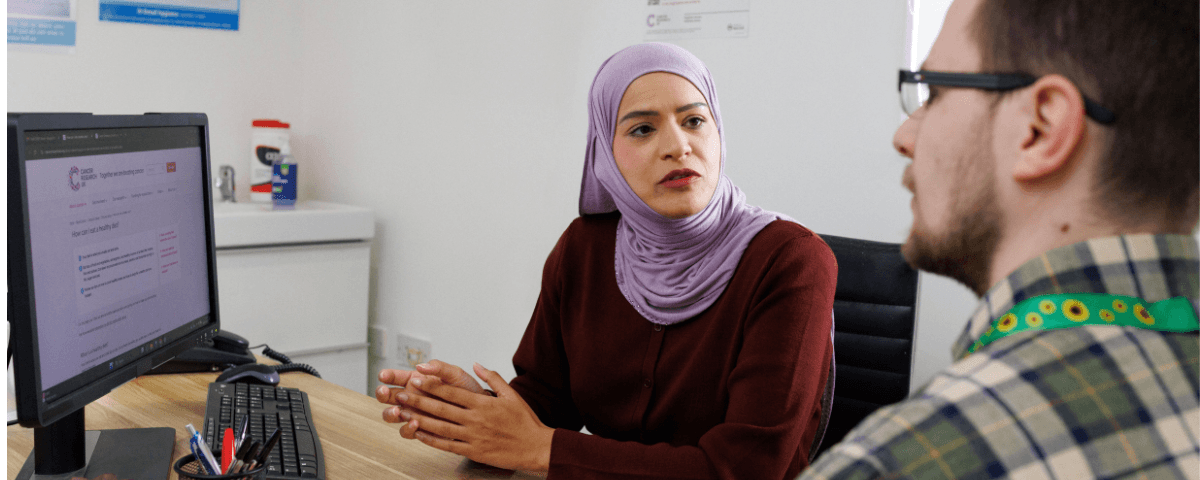 A patient wearing a sunflower lanyard talks to a health professional who is showing them the Cancer Research UK website.