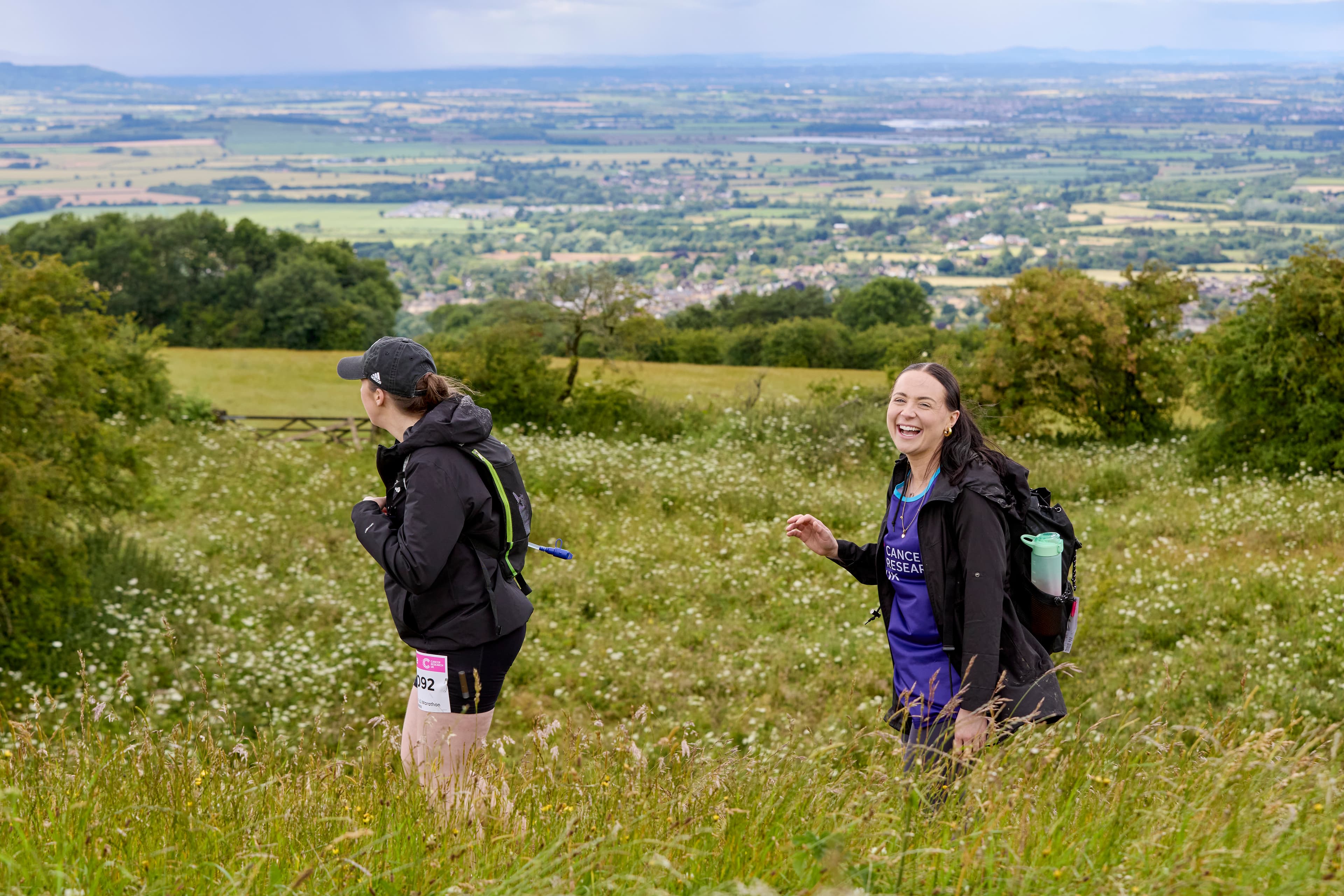 Two Big Hike participants stand on a grassy hill, both wearing backpacks; the person on the right smiles to the camera.