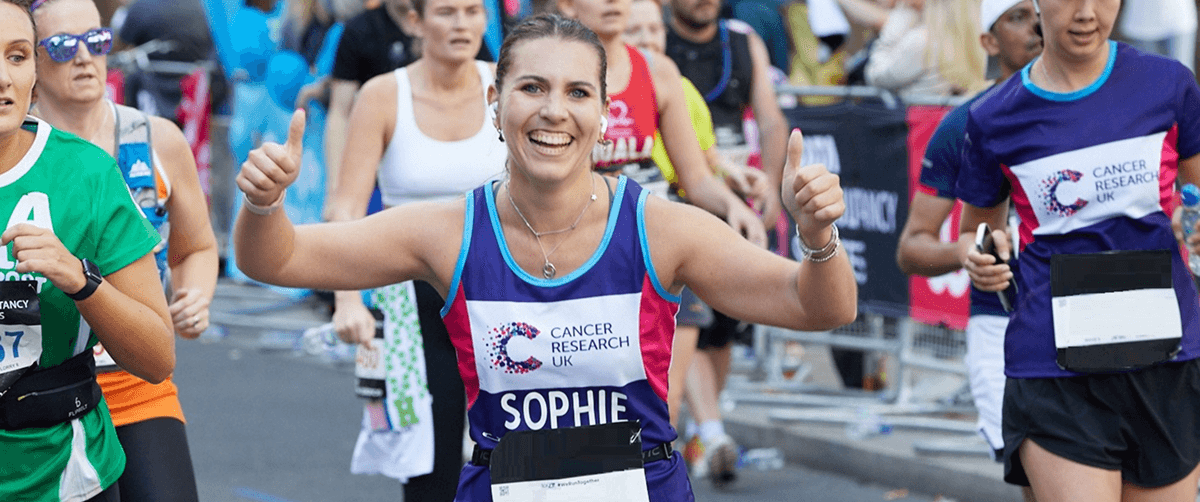 A smilling CRUK Sports runner with the name SOPHIE gives two thumbs up while running a race.